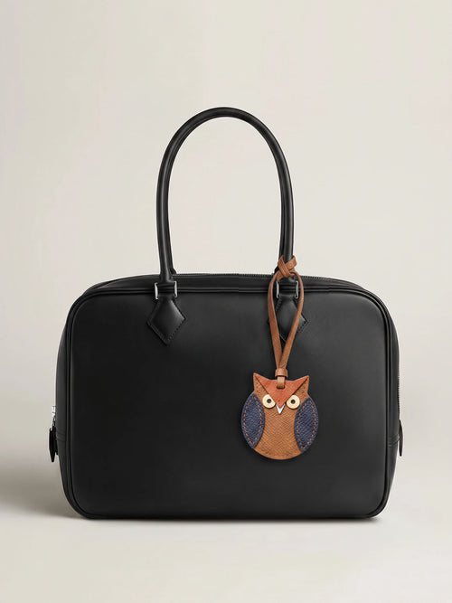 A black leather handbag with a top handle and a hanging owl-shaped charm, photographed against a plain light beige studio background.