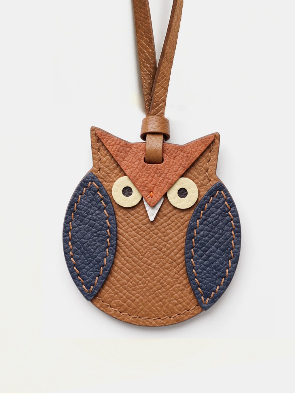 An owl-shaped leather keychain or bag charm in warm brown and tan, with dark navy textured wings and light wood-tone eyes, shown on a plain light background.