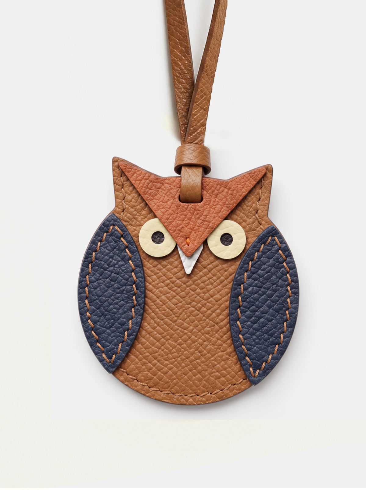 An owl-shaped leather keychain or bag charm in warm brown and tan, with dark navy textured wings and light wood-tone eyes, shown on a plain light background.
