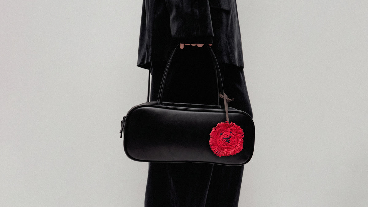 A person in a black outfit holds a black leather cylindrical shoulder bag with a bright red textured lion head charm 