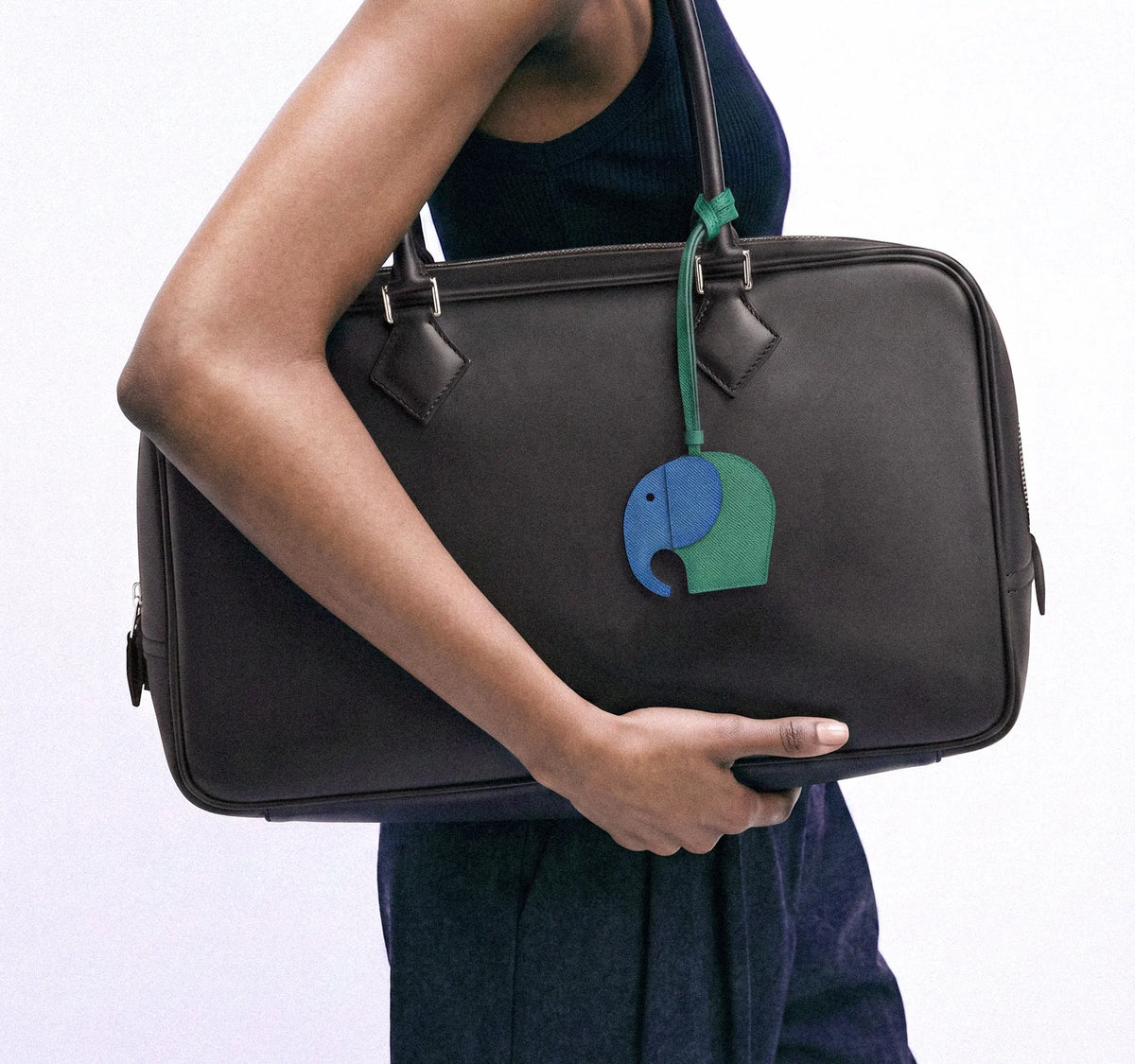 A person holding a large black leather briefcase or duffel bag with a green and blue elephant-shaped hanging charm on the front, against a plain light background.