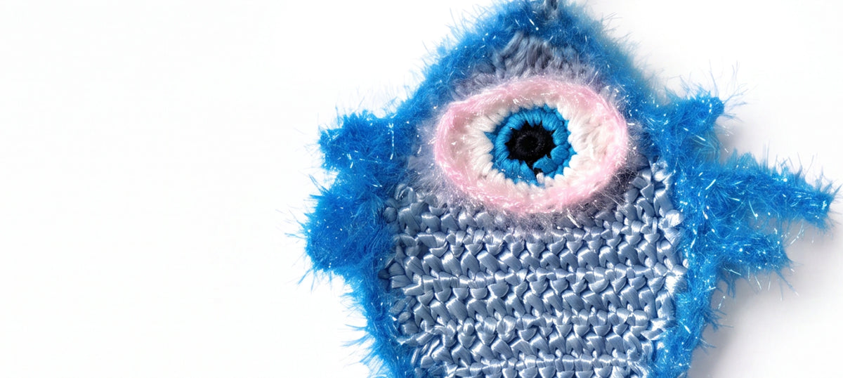 A close-up of a handmade, fuzzy blue fish charm with a single large eye featuring blue, pink, and black details, set against a plain white background.
