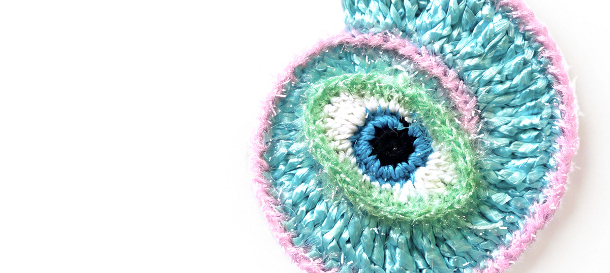 A close-up of a crocheted or knitted circular textile with concentric eye-like rings in blue, teal, mint green, white, and pink on a plain white background.