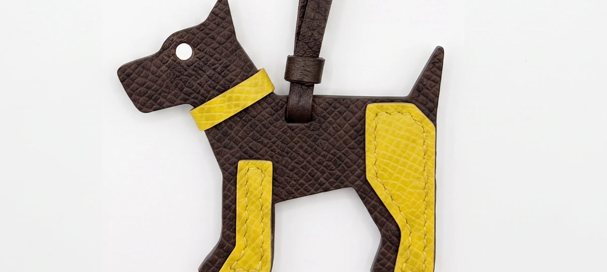 A dog leather luggage tag or bag charm in dark brown textured leather with bright yellow accents on the collar, belly, and hind leg, shown on a plain white background.