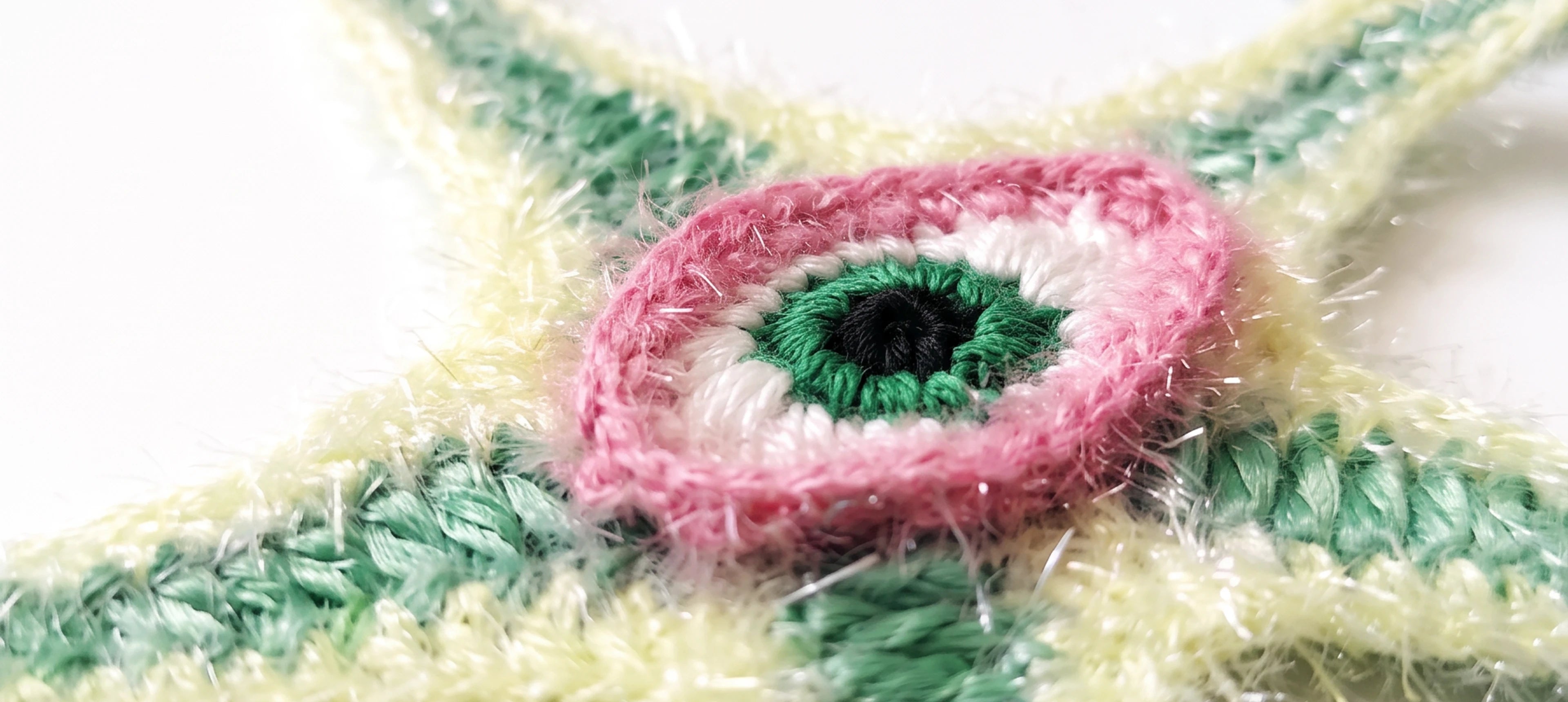 Close-up of a raffia starfish charm with pale yellow arms, green textured stripes, and a central pink, white, and green eye motif on a white background.