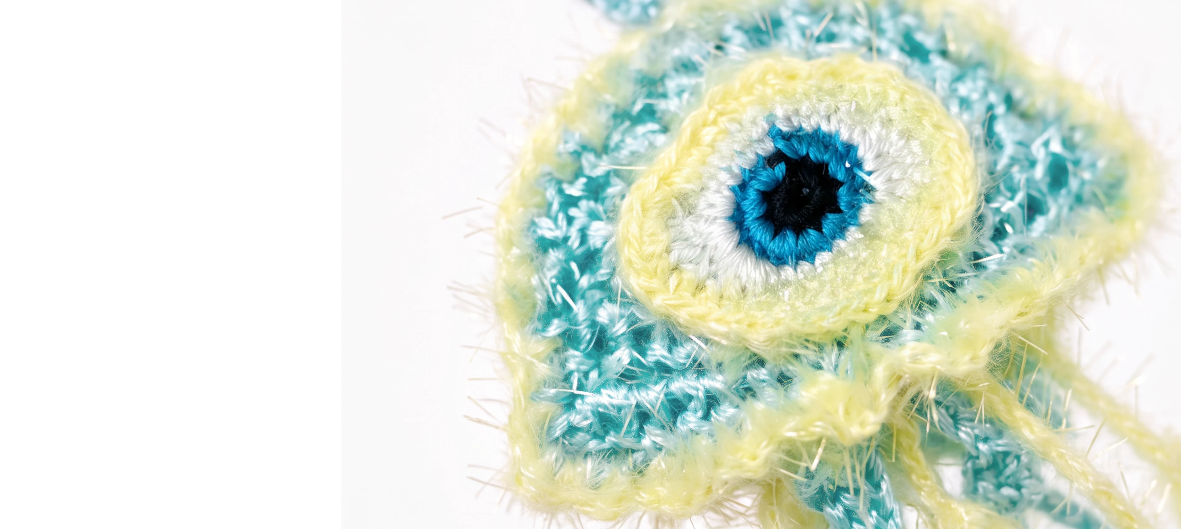 A close-up of a handmade crocheted or knitted eye-shaped accessory in pastel aqua blue and pale yellow yarn, with a black and white center, on a plain white background.