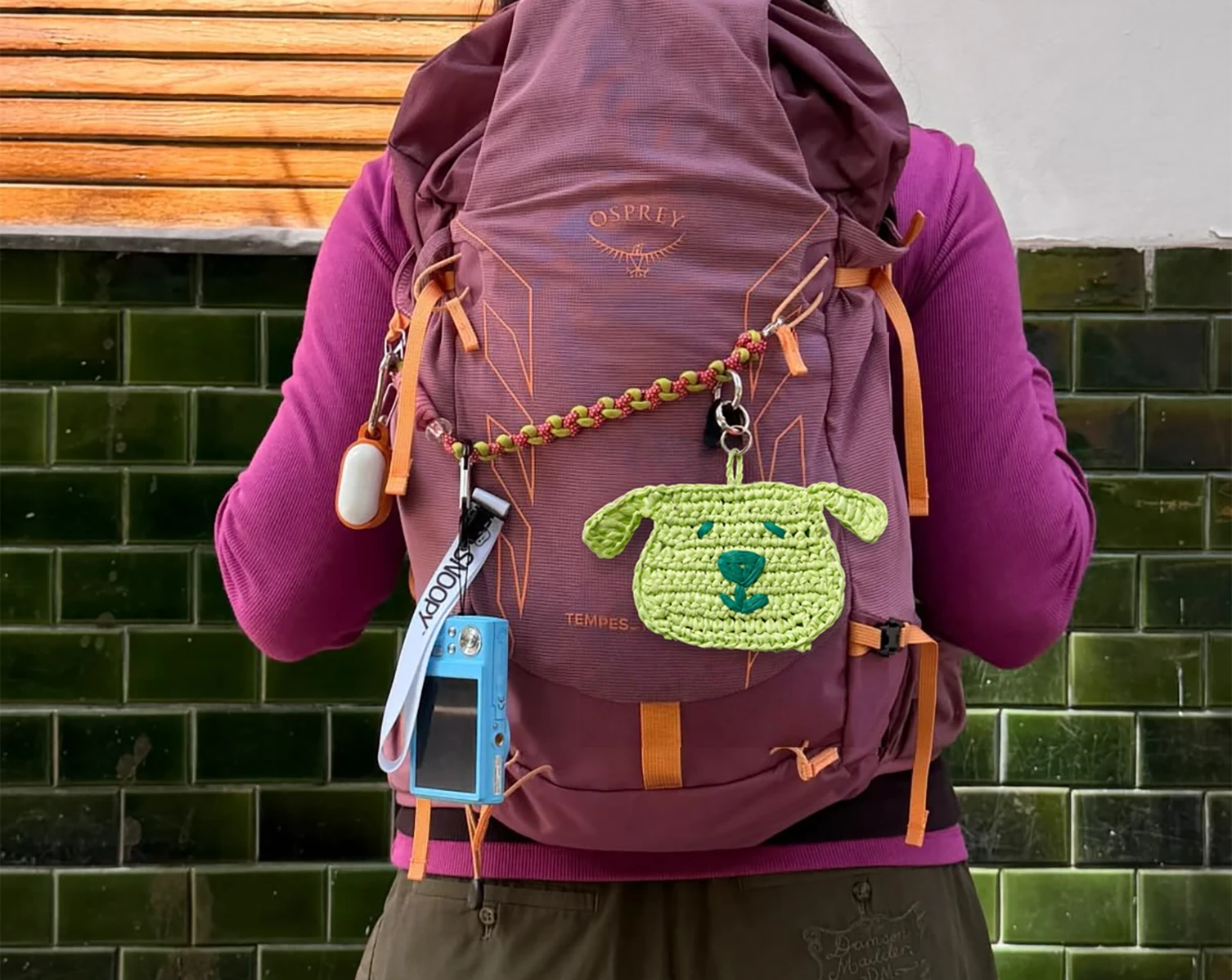 A person wearing a purple backpack with a green knitted dog charm attached.