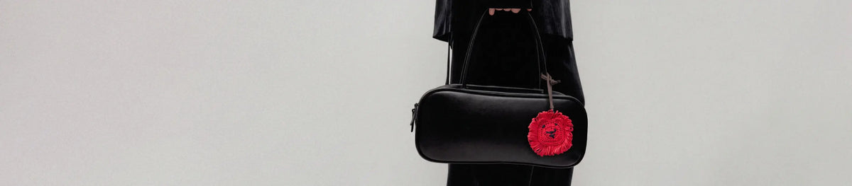 A black leather shoulder bag with a prominent red raffia lion charm, shown against a plain light gray background.