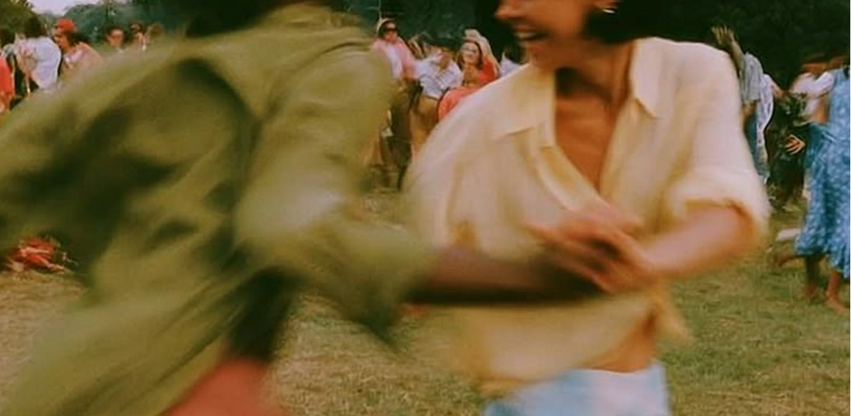 A low-resolution, motion-blurred scene showing two people outdoors on grass, with one person in a light-colored shirt and another in an olive-green top, surrounded by a crowd in the background.
