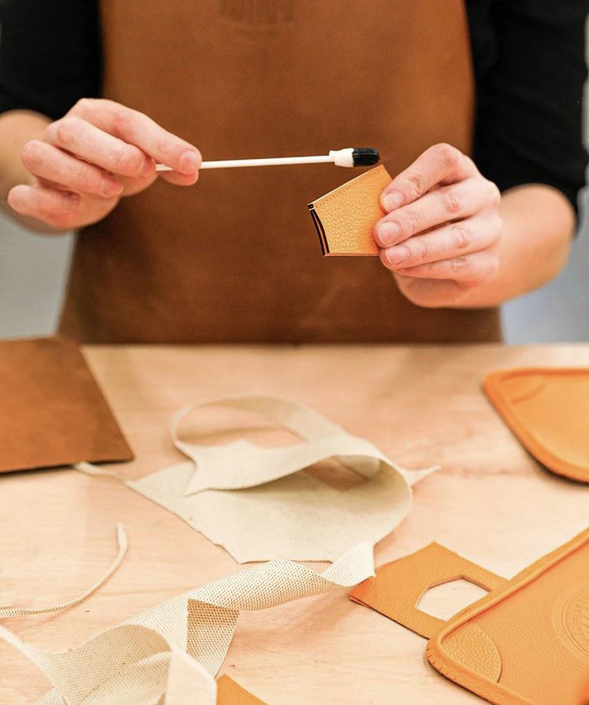 A person wearing a brown apron holds a small tan leather item while applying a black tip to a leather loop with a wooden stick. The scene is set on a light wood table with leather and canvas material pieces scattered around.