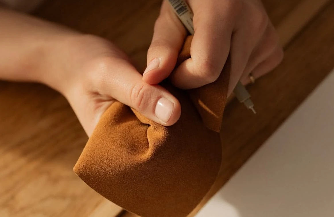 A close-up of a person's hands holding a brown suede or leather pouch while drawing with a gray pen on a wooden surface.