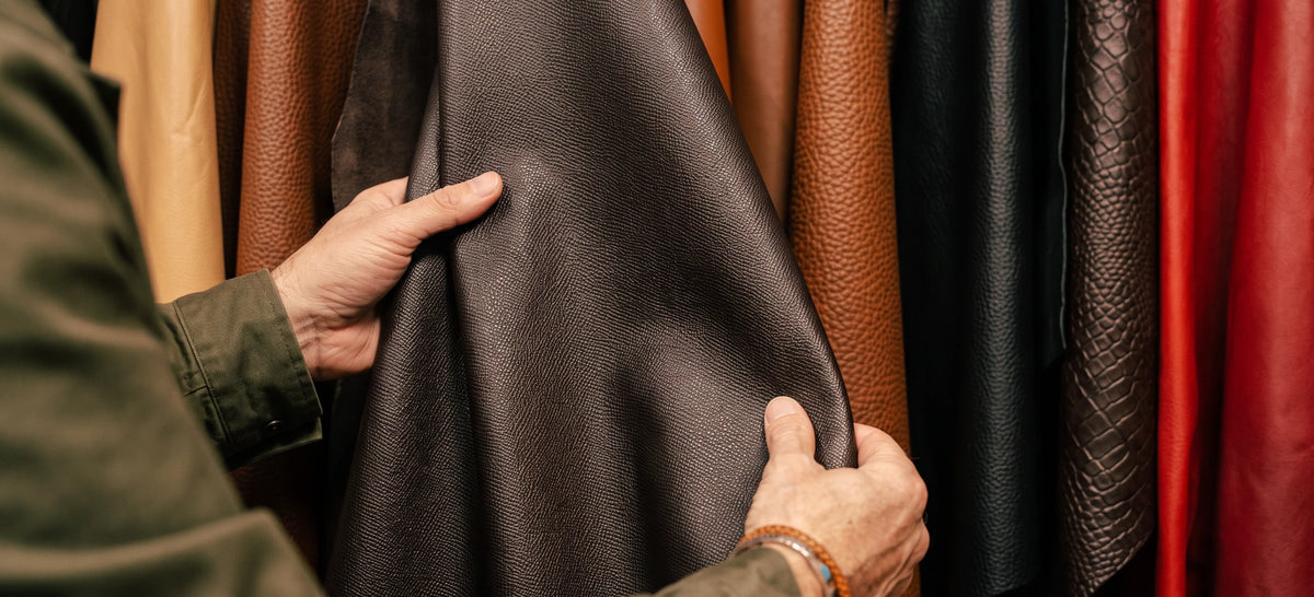 A close-up of a person's hands holding up a dark brown textured leather sheet to show its texture, with other hanging leather rolls in brown, black, and red visible in the background.