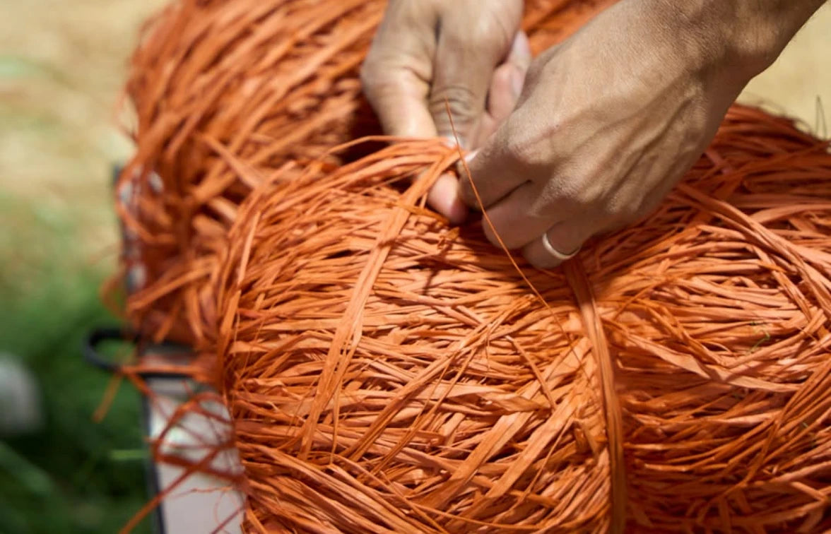 Close-up of a person's hands wrapping orange raffia or straw strips around a bundle, with more orange material visible in the background.