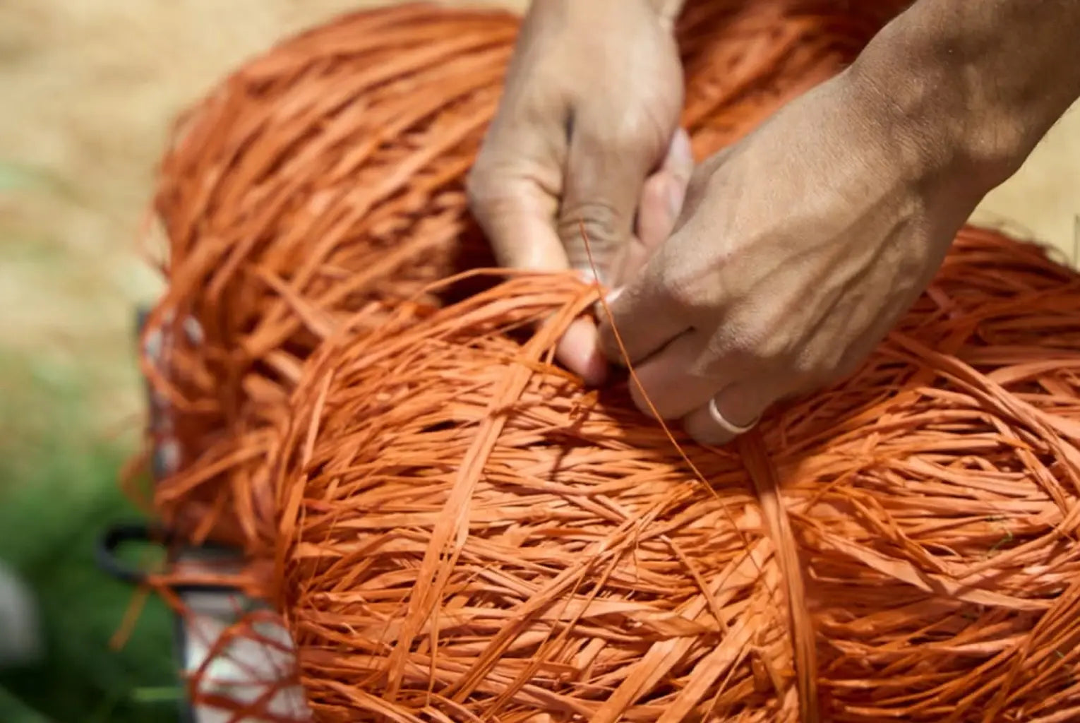 Close-up of a person's hands weaving or binding a bundle of bright orange raffia or straw strips outdoors.