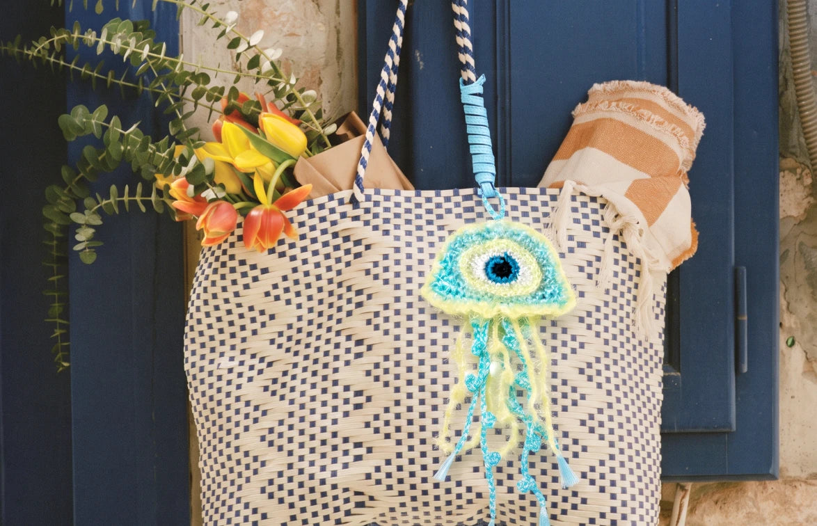 A woven tote bag featuring a woven raffia jellyfish charm with an evil eye in light blue and green.