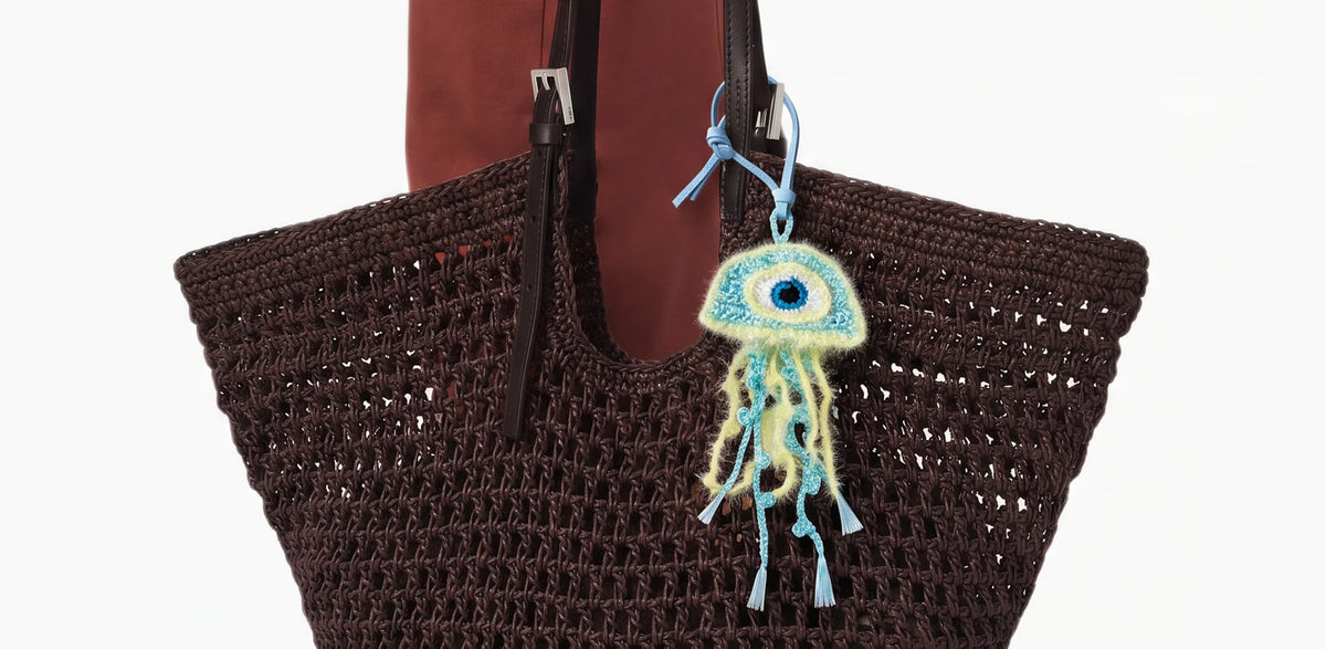 A dark brown crocheted tote bag, featuring a light blue and mint green crocheted jellyfish charm and keychain hanging from one handle against a plain white background.