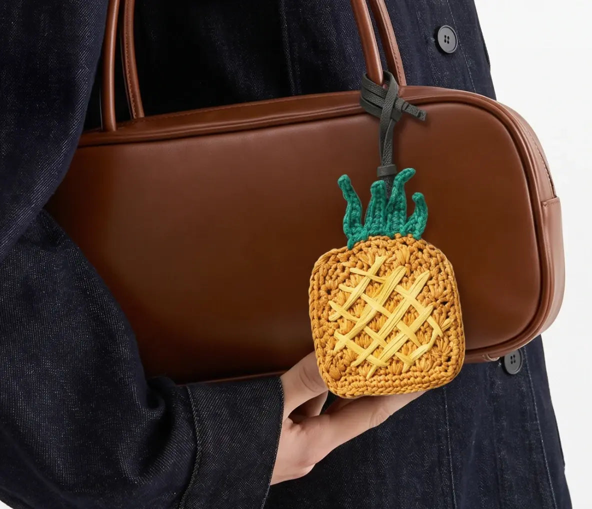 A person holds a brown leather shoulder bag with a small crocheted pineapple bag charm in front of it.