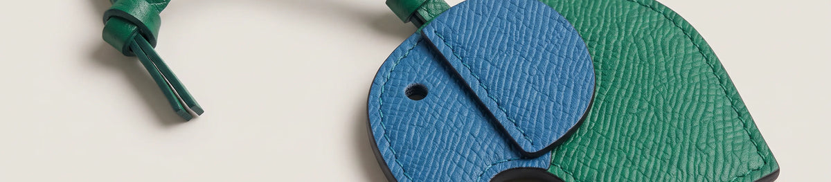 A close-up of a textured leather luggage tag shaped like an elephant, featuring a blue trunk and ear and a green body, on a light beige background.