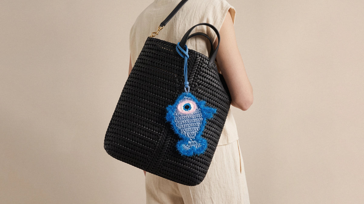 A person carries a black woven straw tote bag with a blue crocheted fish charm hanging from the strap.