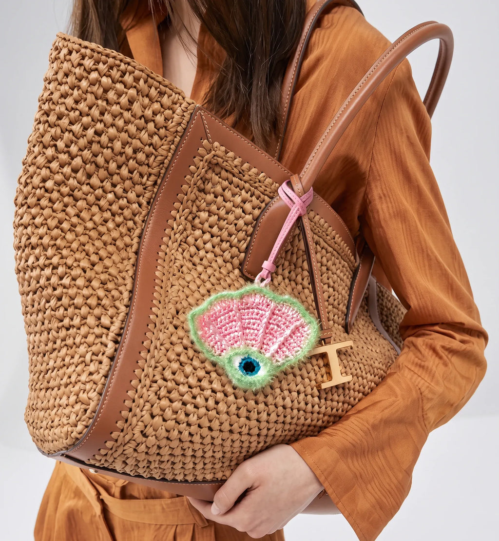 A person wearing an orange-brown satin dress carries a large woven straw tote bag with brown leather trim and handles. The bag features a pink and green embroidered shell charm with a blue eye detail and a small wooden letter T.