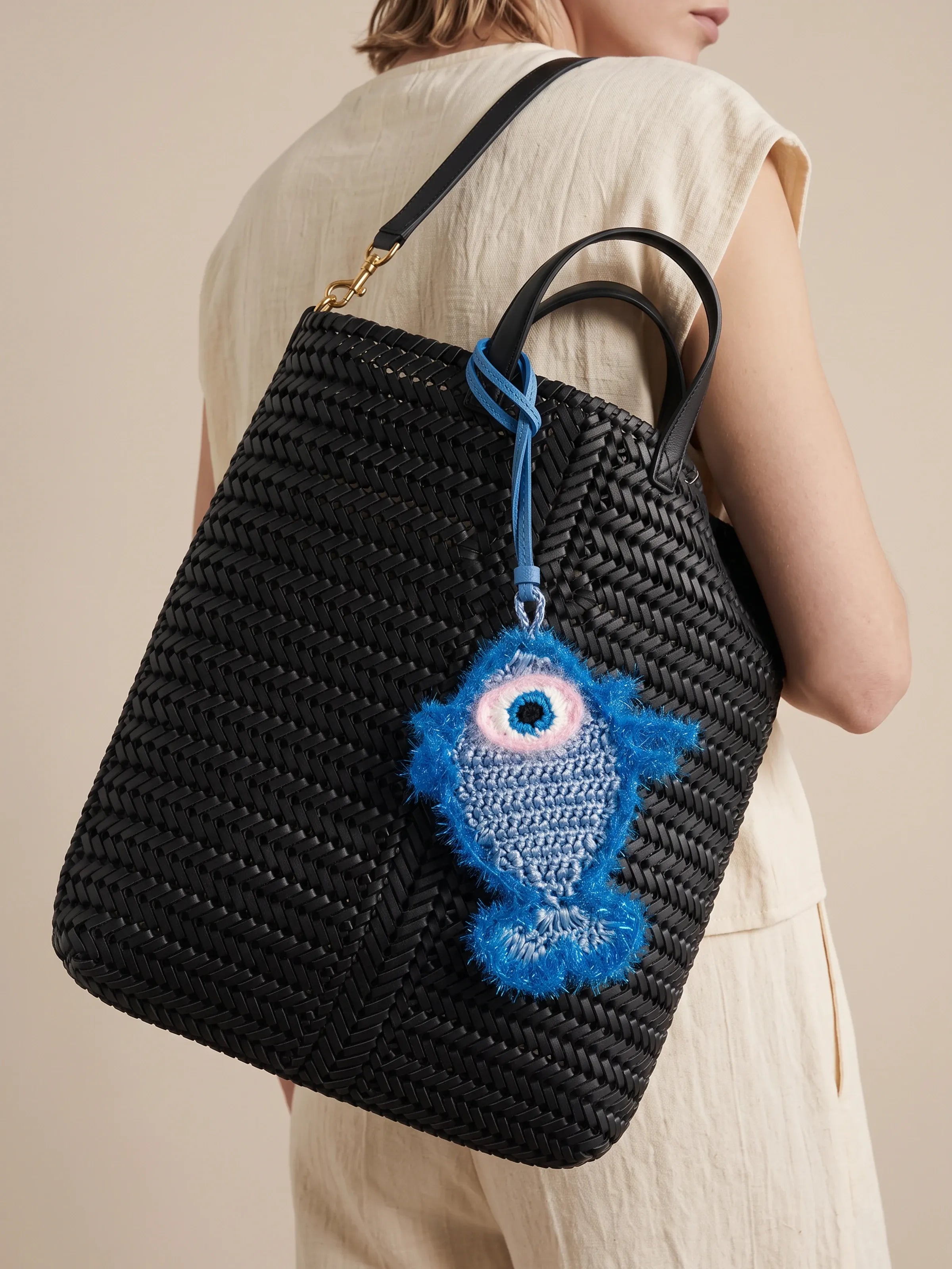 A black woven straw tote bag with a blue knitted fish charm, shown on a model wearing a beige dress against a warm beige background.