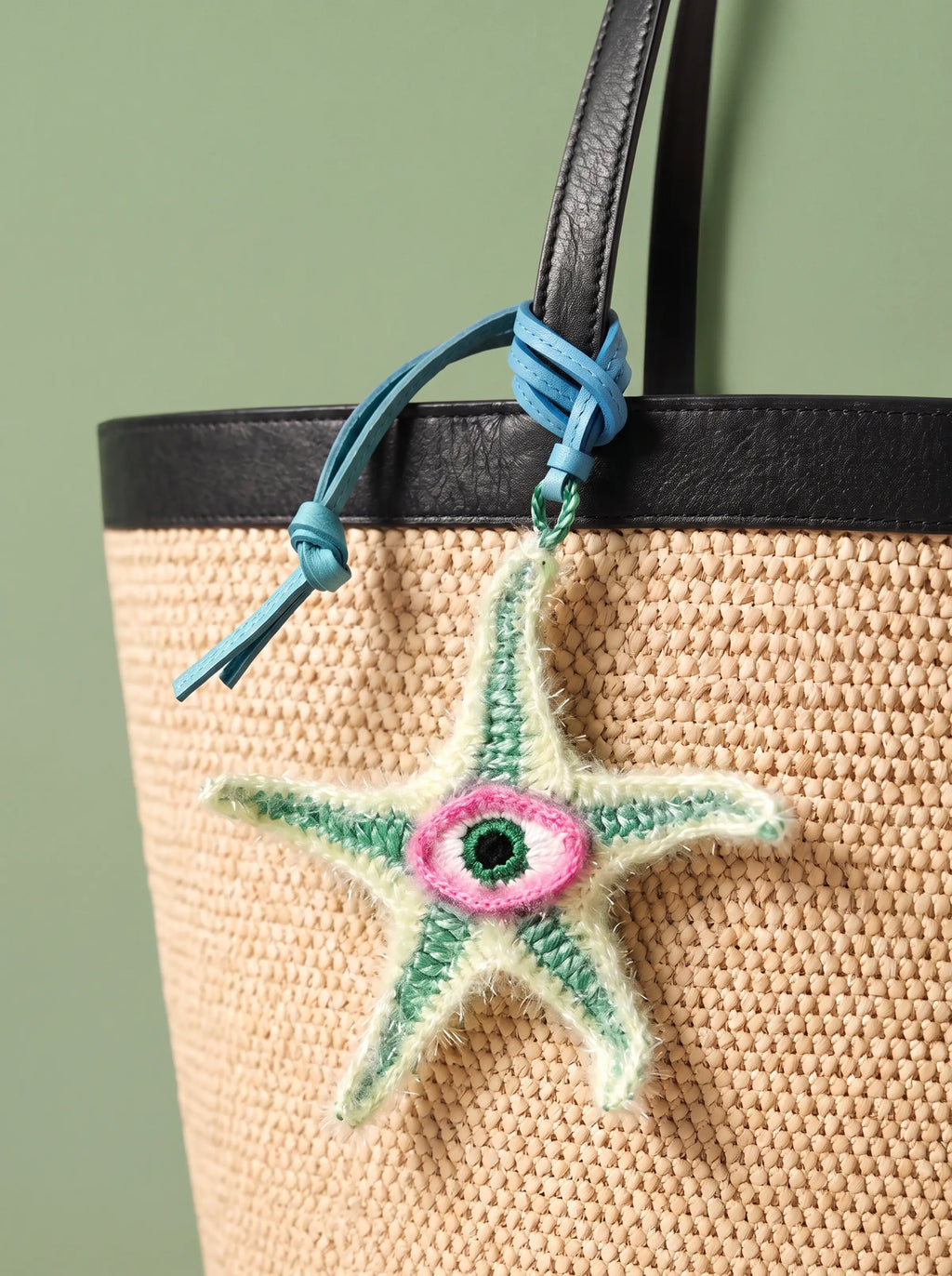 A woven straw tote bag with a black leather trim and handle, featuring a hanging crocheted starfish charm with a pink and green eye design and a blue tie.